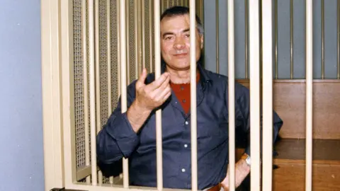 Alamy Mafia boss Nitto Santapaola during his trial for the assassinations of anti-mafia prosecutors. A man wearing a blue shirt is behind white bars and snaking his hand through the bars, with his other hand on his hips