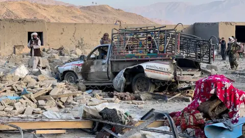 AFP via Getty Images A heavily damaged vehicle with colourful lights installed on the sides is surrounded by rubble from a destroyed building. People stand around it inspecting the area. Mountains are in the distance.
