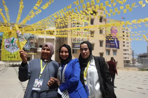 BBC Three Palestinian women stand together, with one holding a yellow Fatah flag, under rows of yellow bunting, in Gaza City