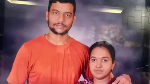 Patnala Bhargavi Patnala Suresh and his wife Bhargavi people pose together. Suresh in an orange shirt has an arm around his wife who is wearing a patterned top, both facing the camera.