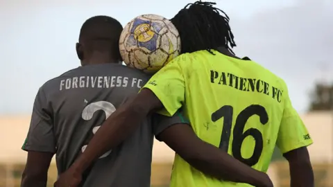 AFP via Getty Images Back view of two football team captains: one from Forgiveness FC (L) - in a black shirt, the other from Patience FC (R) - in a green shirt. They pose a football between their heads after playing a peace football match. Archive shot 2021.