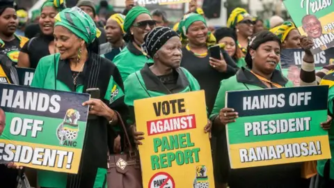 Gallo Images via Getty Images Three ANC Women's League supporters in party colours stand in front of a crowd holding up placards that say 'hands off President Ramaphosa' and 'vote against panel report'.
