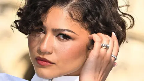 Getty Images Zendaya sporting some silver jewellery and a simple gold band on her wedding finger. She is wearing a crisp white shirt with high collar.