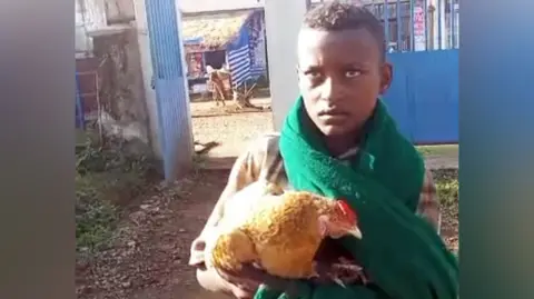 Umer Chane Markos Abaye wears a green scarf and holds a brown chicken. He has a concerned look on his face
