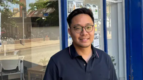 Robbie Kalus/BBC Ray Yu, a young man with short, dark hair, glasses and wearing a blue polo shirt, standing outside a shop.