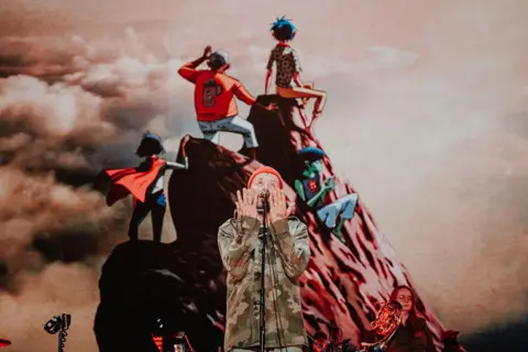 Luke Dyson Damon Albarn sings with his hands raised to his face. In the background, a video screen shows an image of Gorillaz band members standing at the peak of a mountain.