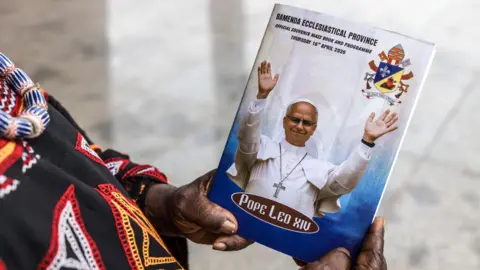 AFP via Getty Images A Catholic worshipper at the Saint Joseph Catherdral in Bamenda, holding a Mass book with the image of the Pope on it. Only the worshippers hands and partial clothing is seen.
