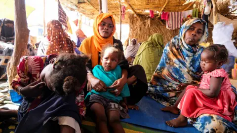 Reuters Two women sit holding two young children. An older child sits to the left of them with her face in her hands. They are in a shelter in a displacement camp after fleeing El-Fasher.