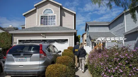 Getty Images A man, wearing an FBI jacket, and a woman in a plain black jacket, walk towards a house with their backs to the camera. The house is two storey, light grey and close to the neighbour. The path is lined either side by manicured bushes, and a silver car is parked on the driveway. 