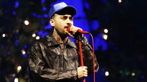Getty Images Zayn Malik wears a leather jacket and a blue hat. He is singing into a red microphone on a red microphone stand.