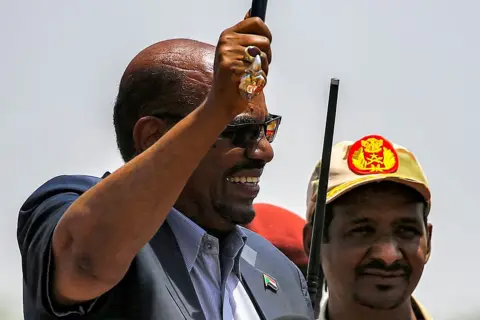 AFP via Getty Images Omar al-Bashir (L) waves a walking stick as he gives a speech at the headquarters of the Rapid Support Forces (RSF) paramilitaries in Umm al-Qura in South Darfur State, while RSF commander Mohamed Hamdan Dagalo, wearing a cap, looks on