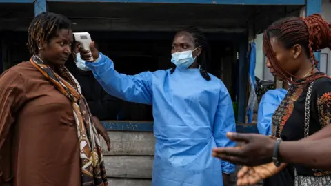Reuters On a street in the DR Congo city of Goma, a health worker in a blue face mask and medical apron takes the temperature of a woman with a device near her head. Another woman and man stand on the other side waiting. 