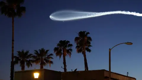 Getty Images The bright white trail of a Falcon 9 rocket carrying 25 Starlink internet satellites is seen against a dark sky, above the silhouettes of a building and palm trees, in Pasadena, California on 6 April 2026 