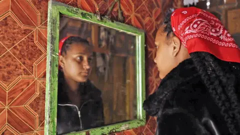BBC A young mixed race woman looks at herself in a mirror - she is wearing a black zip-up top and has braided long hair