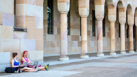 Universal Images Group via Getty Images Two students sitting on the ground with laptops in front of a large building.