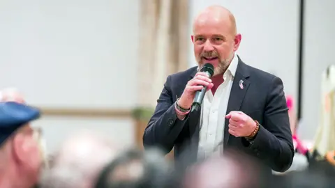 Orange County Register via Getty Images Steve Hilton speaks at a campaign event. He holds a microphone, and wears a black blazer with a pin in the shape of California.