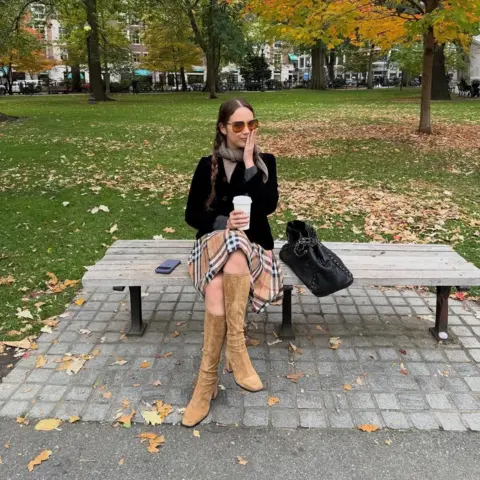 Laufey Laufey sits on a park bench in Boston. She's holding a cup of coffee, with her iphone placed beside her. Her hand is held to her face, as if in contemplation.