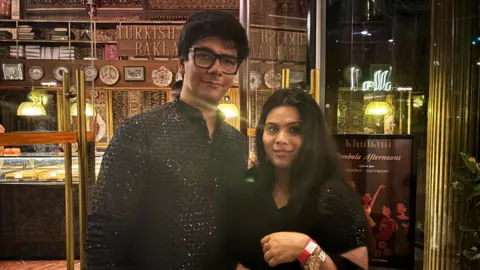 A man wearing black glasses and a black shimmery kurta stands with a woman who has her hair open and is wearing a black Indian suit.