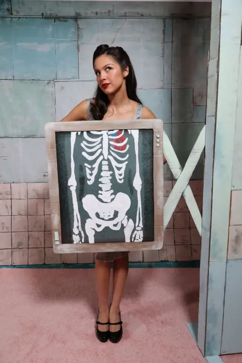 Getty Images Olivia Rodrigo is pictured standing behind a mock-up X-Ray, in a promotional stunt for her single The Cure.