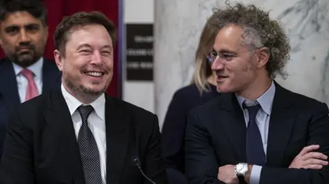 Getty Images Elon Musk and Alex Karp sitting next to each other. Both wear dark suits and ties and are laughing.