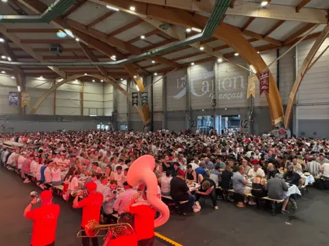 Rows and rows of long tables packed full of people in a large hall with very high ceilings. Painted on the wall at the back of the room are the words Le Canon Français. At the front of the photo there is a brass band standing including a tuba and a trumpet and two cornets. They are wearing red T-shirts and red baseball caps.