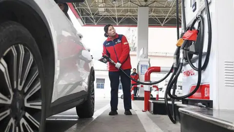 Getty Images A woman in a red jacket looks at the metre on the petrol pump as she holds the hose near the rear of a white car