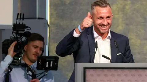 Reuters Ulrich Siegmund, who has a dark blonde undercut which is swept back on the top, grins at his audience from a lectern and does a thumbs up 