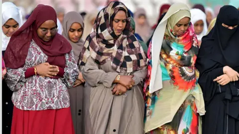 AFP via Getty Images A group of women in colourful head scarves stand and bow their heads and clasp their hands during Eid prayers in Port Sudan.