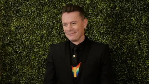 Bloomberg via Getty Images Larry Mullen Junior, a man with fair hair, wearing a dark suit. There is a green hedge in the background.