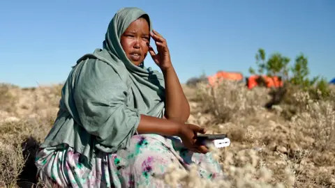 BBC/Amensisa Ifa A woman sitting on a mountainside wearing a light grey scarf covering her head and shoulders. She is looking into the distance and holding a mobile phone on her lap.