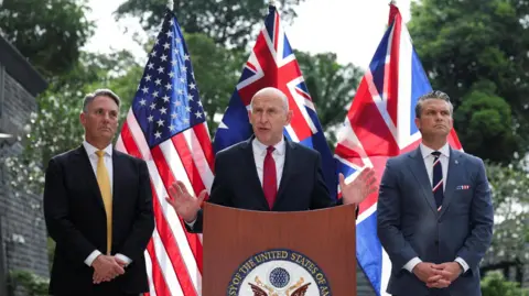 Reuters John Healey speaks and gestures with his hands at a podium at the US embassy, flanked by Richard Marles and Pete Hegseth. The US, UK and Australia flags are posted behind them.