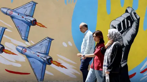 Getty Images An older man and woman and a younger woman walking past a mural in Tehran featuring several drones and a raised fist