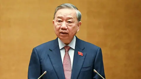 AFP via Getty Images To Lam wears a dark suit, a white shift and maroon tie while speaking at a lectern