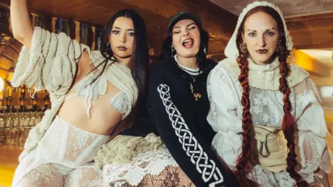 Sarah Ellis Three women pose together inside a warmly lit bar with wooden walls and shelves of bottles in the background, wearing layered and textured outfits. Two of them have brown hair, and the third has long red plaits with a hood.