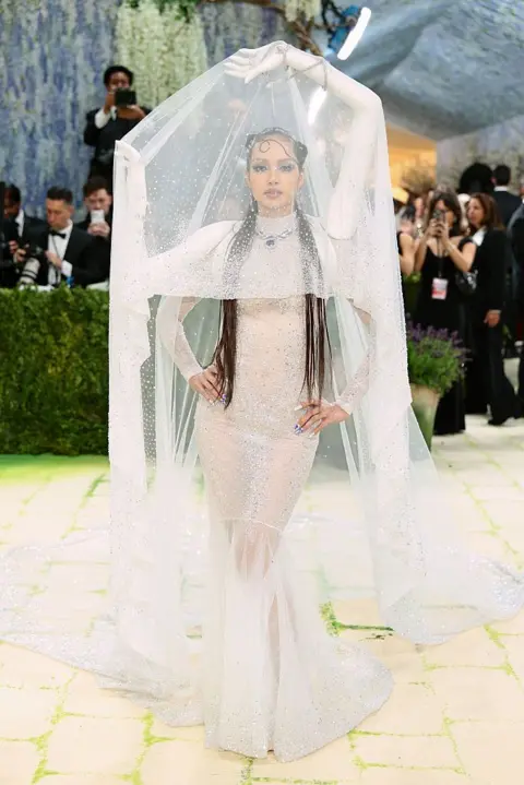 Getty Images Lisa, a member of the K-Pop group BLACKPINK, wears a white gown with an interesting illusion: mannequin arms hold up a white veil, while her real arms are free to pose.