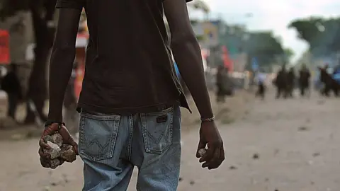 AFP via Getty Images A man seen from behind walking down a street in Nairobi, holding several rocks in their hands, with a blurred crowd visible in the distance