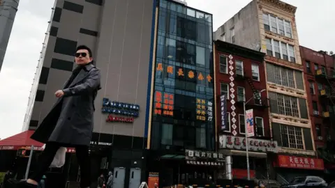 Getty Images A man in a black trench-coat walks in front of a row of buildings in Manhattan's Chinatown