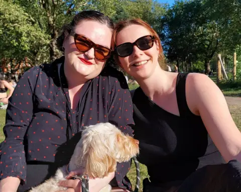 Two women in black tops and sunglasses sit, smiling, in a park with a dog on their lap