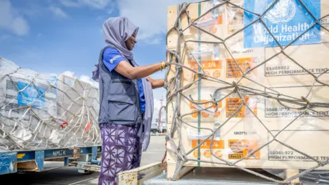 World Health Organization/Handout via Reuters A woman inspects large boxes of aid on shipment pallets