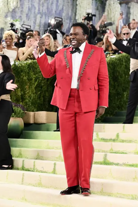 Getty Images Broadway actor Joshua Henry wears a red double-breasted tuxedo with silver lapel embellishments. He stands on the stairs and sings as he performs for the press.