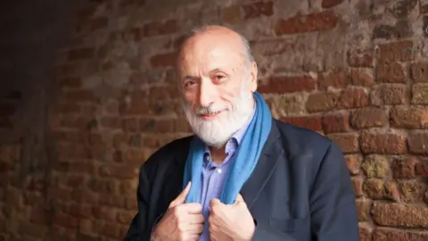 Getty Images Smiling Carlo Petrini poses in front of an red brick wall. He is wearing a dark grey suit jacket and light purple shirt and is clutching a blue scarf with both hands that is around his neck.