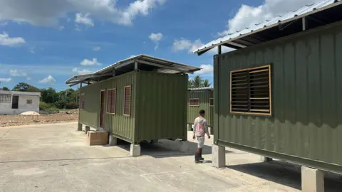 A man is seen from behind walking between two green containers with windows, doors and roofs. 