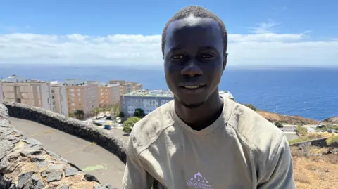 BBC A young Gambian man smiles to the camera in a t-shirt. Behind him is the coastline and apartment buildings 