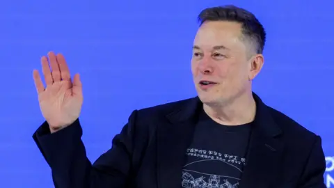 Reuters Elon Musk waving to a crowd from a stage wearing a black blazer and a black t-shirt.