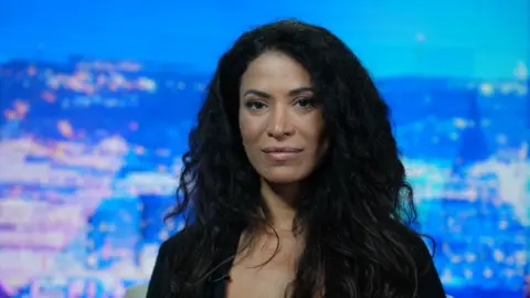 Thierry Humeau Lisa Phillips with wavy black hair, looks straight ahead. She wears a black top. A blue and purple cityscape sits in the background.