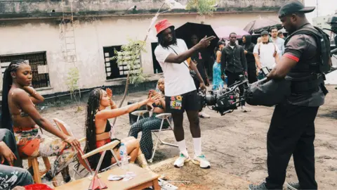 Meji Alabi Meji Alabi in a red hat, white T-shirt, black shorts and white socks and trainers holds a camera and points during a shoot. To his side are women sitting on deck chairs by a spindly tree. A cameraman, with a black cap and dressed in dark clothes, stands to his other side and production staff, some holding black umbrellas, stand behind.