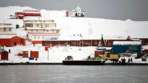 Getty Images A series of single storey building coloured brown and orange stand in the snow comprising Chile's Presidente Eduardo Frei military base next to the Antarctic Ocean. People can be seen working along the shoreline and there is an open-topped boat in the water. 