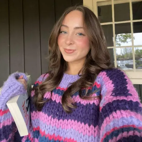 Meagan Carioti Picture of Meagan in a purple jumper, holding a book.