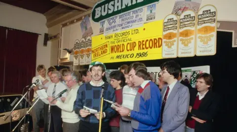 Getty Images Northern Ireland football team members, including Gerry Armstrong in green and white cap dark hair and moustache and checked jumper. Manager Billy Bingham is in a white jumper and glasses. Pat Jennings is in blue, grey and red jumper.