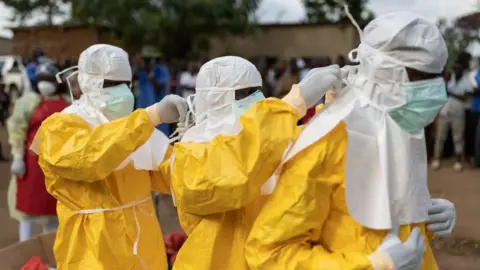 Getty Images The image shows three individuals in the foreground wearing full-body protective suits in bright yellow, along with white hoods, face masks, and gloves. Two of the individuals are helping adjust or fasten the protective gear of a third person, focusing on securing the hood or mask around the head and neck area. The setting is outdoors, with a blurred crowd of people and buildings visible in the background, suggesting a public or community environment. Additional people in the background are also wearing protective clothing. The lighting is natural daylight, and the scene emphasizes careful preparation and attention to safety procedures.